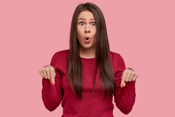 Portrait of attractive young woman has surprised face and opened mouth, points down at floor, wears casual clothes, models against pink background, demonstrates dirty shoes. Emotions concept
