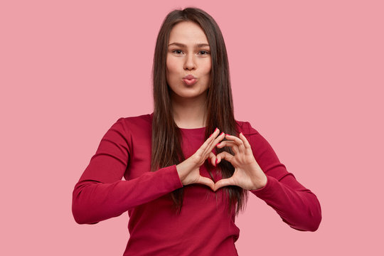 Photo of pleasant looking woman makes lips round, shows heart gesture with fingers, flirts with boyfriend on distance, has appealing appearance, wears red clothing, models over pink background