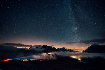 Long distance vehicle lights coming in the left side. Beautiful space filled with stars in the sky. The mountains are surrounded by dense fog
