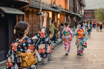 Japanese girl in kimono taking a photo of a traditional street with wooden houses on her cell phone in Kanazawa Japan - obrazy, fototapety, plakaty