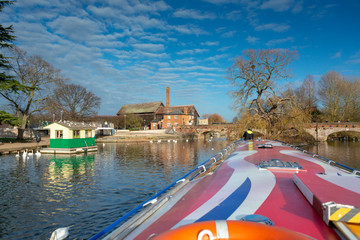 man stands on front of boat in bright sunlight during winter in the UK with ancient buildings and bridges in the background