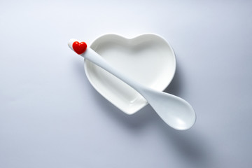 Concept: love with the first spoon for Valentine's Day or wedding romantic menu. Ceramic plate in the form of a heart and a spoon with a red symbol of love on a white background. Minimalism. Close-up.