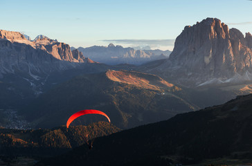 Paraglider with red parachute flies over the mountains filled with trees in the sunlight
