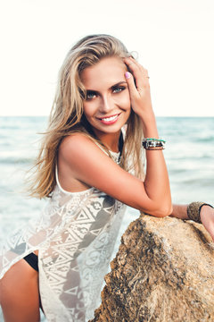Attractive blonde girl with long hair  is posing to the camera on sea background. She wears black bikini under white dress. She leans on stone and looks to the camera.