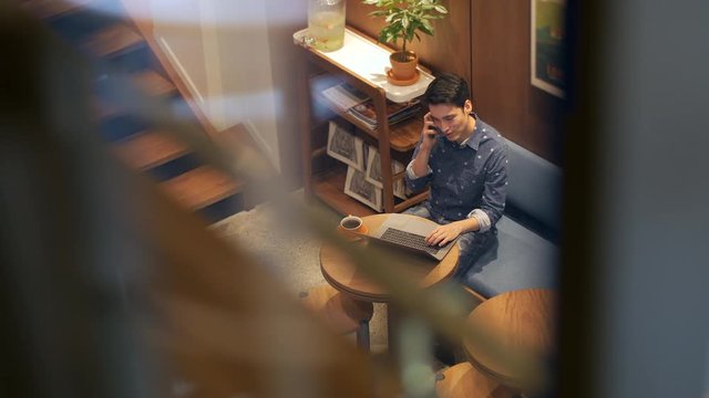 Hispanic Latin American young man "digital nomad" sitting in coffee shop with laptop talking on cell phone, top view through foreground