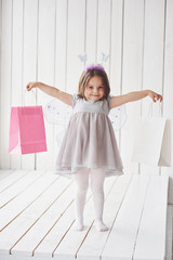 Holding paper packages while standing on the white wooden floor. Beautiful little girl with fairy costume having fun posing for the pictures