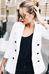 Portrait beautiful woman in sunglasses on street . She wears white jacket, touching hair, looking down