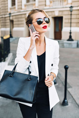 Portrait attractive woman in sunglasses with red lips walking on street . She wears white jacket,...
