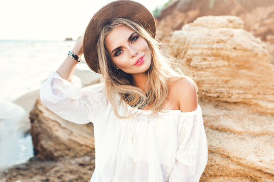 Portrait of attractive blonde girl with long hair posing on deserted beach. She wears white shirt, hat, ornamentation. She is smiling to the camera.