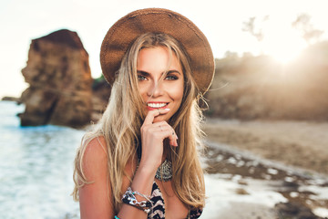 Closeup portrait of attractive blonde girl with long hair posing on rocky beach on sunset background. She wears bikini, hat. She holds finger on lips and smiles to the camera.