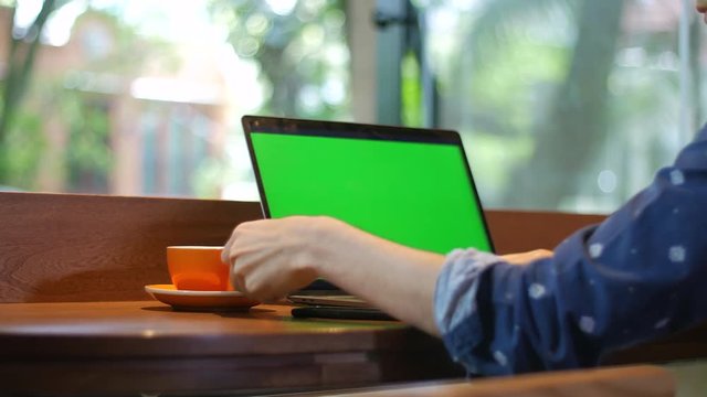 Close-up of laptop computer with chroma key greenscreen, with hands of a man typing and drinking coffee from a cup, with view outside from coffee shop