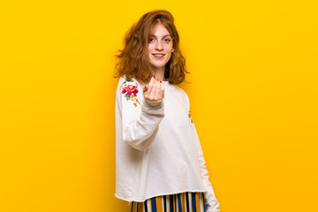 Young redhead woman over yellow wall inviting to come with hand. Happy that you came