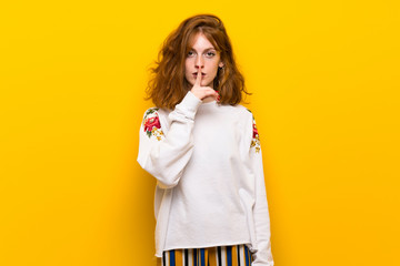 Young redhead woman over yellow wall showing a sign of silence gesture putting finger in mouth