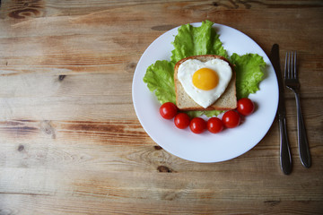 fried eggs with a yellow yolk in the form of a heart on bread, decorated with green salad and red cherry tomatoes on a white plate and wooden rustic background with space for text 