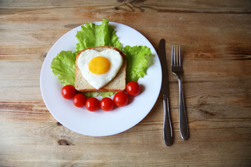 fried eggs with a yellow yolk in the form of a heart on bread, decorated with green salad and red cherry tomatoes on a white plate and wooden rustic background with space for text 