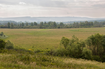 Grey sky with a lot of clouds in the early autumn sky over green fields, trees, forests and huge mountains. A lot of meadow herbs around. Day. Travel through nature landscape