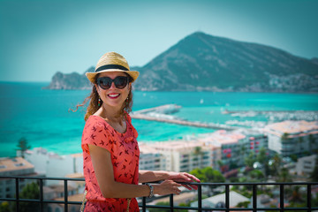 Fototapeta na wymiar Happy tourist woman with straw sunhat looking to the mediterranean sea and enjoying the blue and scenic seascape in Altea, Alicante, Spain. Living coral dress, color of the year