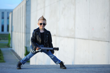 A  little, cute boy guitarist in leather jacket and sunglasses  playing guitar.  Young idol. Children's interests and hobbies. Be like a rock star.