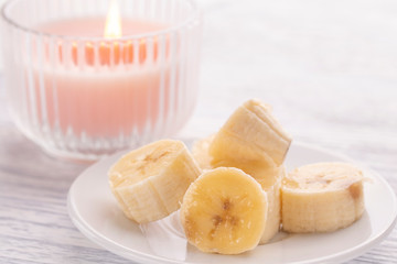 Sliced banana on a white plate and a light wooden table. Pink burning candle nearby.