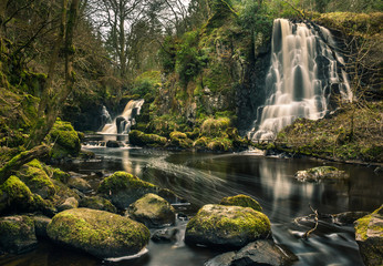 Long exposure shot of Linn Jaw Waterfalls, near Livingston, Scotland, with mossy rocks in the foreground and surrounding the waterfalls and white foam streaks in the water. West Lothian. UK. Nature. L