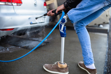 Young man with prosthetic leg washing his car using high pressure water. Selective focus.