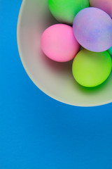 Overhead view of brightly painted easter eggs in a white bowl on blue background