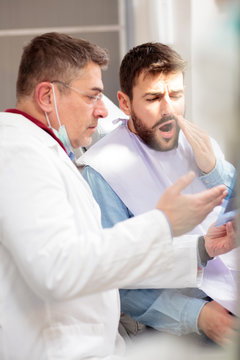 Serious mature male dentist showing x-ray image to a young male patient with toothache. Dental exam in medical clinic