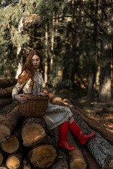 Young woman with long red hair in a linen dress gathering mushrooms in the forest in the bast basket