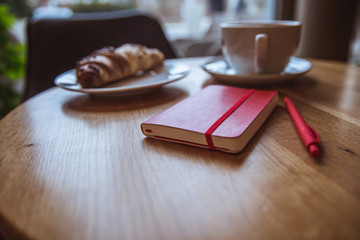 A red note and red pen is on the table in a cafe, a cup of coffee and a croissant on background