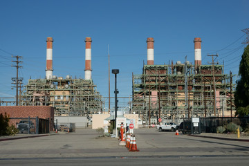 The entrance is shown of a modern power generating station with four tall, red and white chimney smoke stacks in the background during the day.