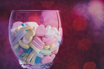 Close up of a wine glass filled with pastel valentine love message candies surrounded by bokeh on a red velvet background