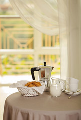 Fototapeta na wymiar Coffee maker, cups and croissants in the basket are on a round table in front of the window.