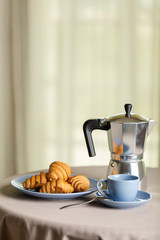 Coffee maker, cups and croissants in the basket are on a round table in front of the window.
