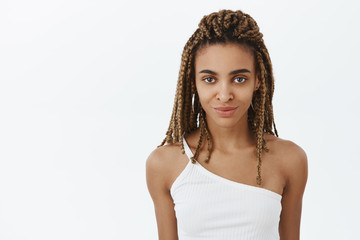 Waist-up shot of confident good-looking delighted dark-skinned woman in top with dreadlocks smiling daring having interesting idea and intention in creative mind against grey wall