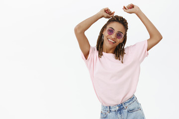 Fototapeta premium Shake that body. Carefree stylish and attractive modern African American female with cool dreads in trendy sunglasses raising hands dancing and smiling enjoying awesome party feeling confident