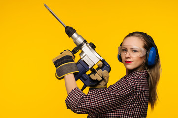 Beautiful young woman in goggles and headphones worth holding perforator drill isolated on yellow background