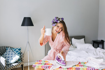 Pretty housewife with long hair  in pink bathrobe with curler on head at home at ironing clothes....