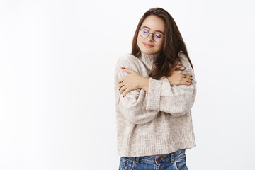 Girl feeling great in soft cozy sweater during chilly weather hugging herself and smiling from comfort and delight close eyes, enjoying warm outfit on cold fall evening over gray background