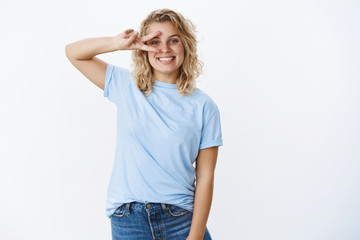 Peace my friend. Portrait of happy and kind sincere cute european female student with blue eyes and short curly hair showing victory or disco gesture on eye and smiling positive, in good mood