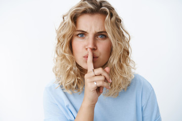 Shush you cannot say it. Portrait of displeased blond girl with short haircut and blue eyes looking with dismay frowning saying shh with index finger on mouth telling keep secret over white wall