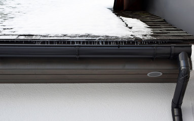 Grey plastic rain gutter with drain downspout pipe installed on asphalt shingles roof, wooden eaves with round ventilation grille and roof covered with snow and icicles in winter day.