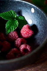 Freshly picked raspberries in a modern dark blue bowl with a fresh green raspberry plant leaf.