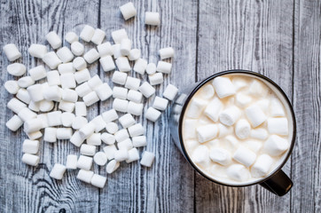 A cup of cocoa with marshmallows, around which marshmallows are scattered on a wooden gray...