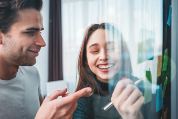 Smiling business man and woman using sticky glass note wall while brainstorming project at space office. Team of coworkers people discussing new startup plan and posted on a sticky glass note wall