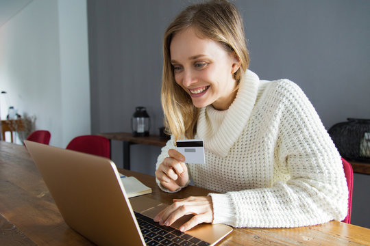 Cheerful businesswoman scanning credit card using web camera of laptop. Positive young woman in white sweater making online purchase. Online shopping concept