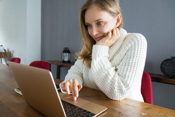 Content pretty woman in white sweater using laptop at home. Young female freelancer analyzing...