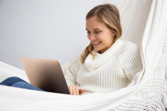 Happy student sitting in hammock and using laptop. Smiling young woman working at home. Technology concept