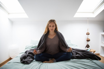 Serene young woman sitting in lotus position on bed and meditating in small bedroom. Serious girl...
