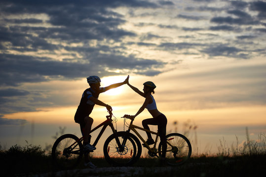 Adorable couple in helmets taking family ride on bicycles, standing and posing. Silhouettes of sporty man and woman highing five. Amazing sunset and beautiful sky background.