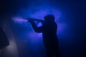 Silhouette of man with assault rifle ready to attack on dark toned foggy background or dangerous bandit in black wearing balaclava and holding gun in hand. Shooting terrorist with weapon theme decor